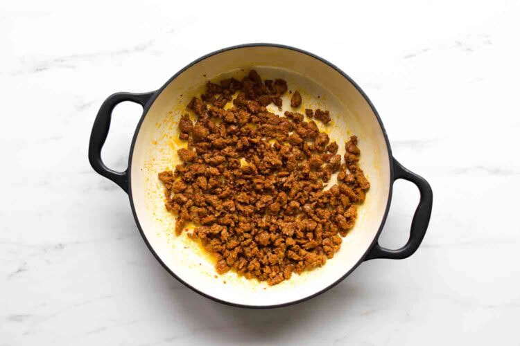 A white pot with black handles contains cooked and crumbled seasoned ground meat, perfect for adding to goat cheese tomato pasta, on a white surface.