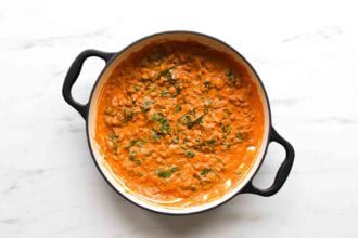 A black pot filled with creamy orange curry, reminiscent of goat cheese tomato pasta, with visible spinach and herbs on a white marble surface.