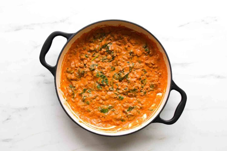 A black pot filled with creamy orange curry, reminiscent of goat cheese tomato pasta, with visible spinach and herbs on a white marble surface.