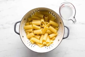 Cooked rigatoni pasta draining in a metal colander, with a glass measuring cup of pasta water next to it—perfect for making creamy goat cheese tomato pasta on a white countertop.