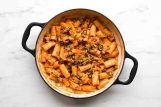Large pot filled with rigatoni pasta in a creamy goat cheese tomato sauce with visible spinach and herbs, set on a white marble surface.