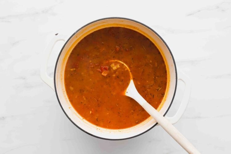 A white pot filled with rich, tomato-based Marry Me Chicken Soup sits on a white surface, with a white spoon resting inside.