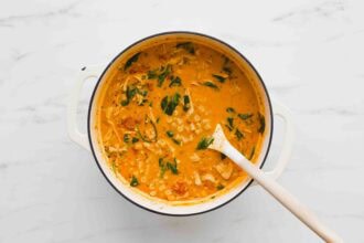 A white pot filled with creamy Marry Me Chicken Soup—tomato soup with pasta, shredded chicken, and spinach—sits on a marble surface with a wooden spoon inside.