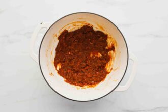 A white pot containing a thick, red-brown tomato-based sauce with visible onion and spice pieces, reminiscent of Marry Me Chicken Soup, set on a white marble surface.