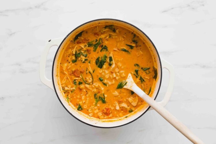 A white pot filled with creamy Marry Me Chicken Soup—tomato soup with pasta, shredded chicken, and spinach—sits on a marble surface with a wooden spoon inside.