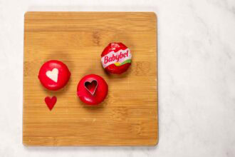Three Babybel cheese rounds on a wooden charcuterie board; two feature heart shapes cut into the red wax coating, with a heart-shaped wax piece beside them—perfect for a Valentine's Day charcuterie board.
