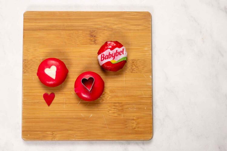 Three Babybel cheese rounds on a wooden charcuterie board; two feature heart shapes cut into the red wax coating, with a heart-shaped wax piece beside them—perfect for a Valentine's Day charcuterie board.