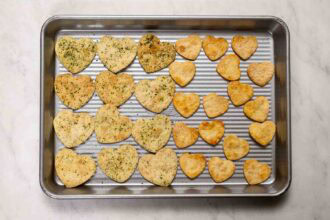A baking tray with heart-shaped crackers, some topped with herbs and some plain, arranged in rows on a marble surface—perfect for adding a festive touch to your Valentine's Day charcuterie board.