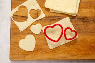 Two heart-shaped cutters and slices of cheese on a wooden cutting board, perfect for adding to your Valentine's Charcuterie Board with adorable heart-shaped cheese cutouts.
