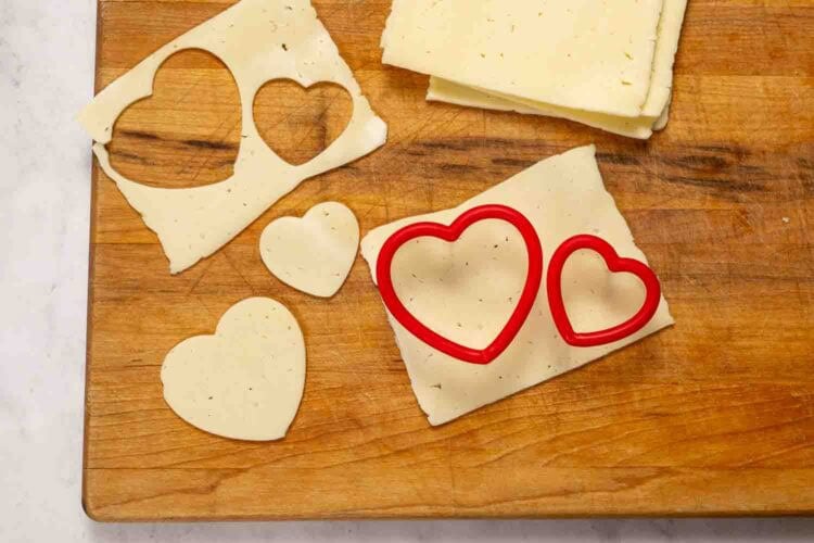 Two heart-shaped cutters and slices of cheese on a wooden cutting board, perfect for adding to your Valentine's Charcuterie Board with adorable heart-shaped cheese cutouts.