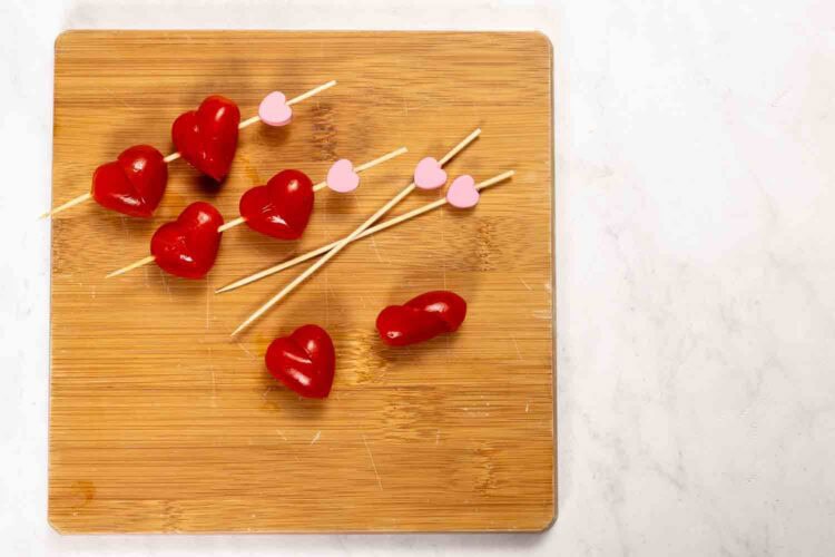 Wooden charcuterie board with red heart-shaped gummy candies, some skewered on sticks with pink heart toppers, perfect for a sweet Valentine's Day treat on a light surface.