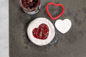 A round wheel of cheese with a heart-shaped cutout filled with red jam, a red heart-shaped cutter, and a jar of jam on a dark surface—perfect for your Valentine's Charcuterie Board.