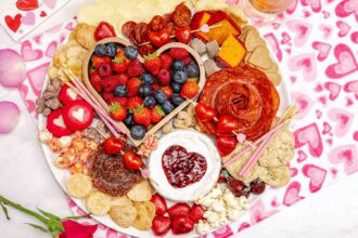 A round Valentine's Charcuterie Board with assorted fruits, cheeses, meats, crackers, cookies, and heart-shaped decorations is set on a pink heart-patterned tablecloth.