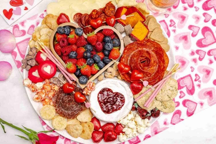 A round Valentine's Charcuterie Board with assorted fruits, cheeses, meats, crackers, cookies, and heart-shaped decorations is set on a pink heart-patterned tablecloth.