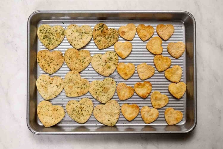 A baking tray with heart-shaped crackers, some topped with herbs and some plain, arranged in rows on a marble surface—perfect for adding a festive touch to your Valentine's Day charcuterie board.