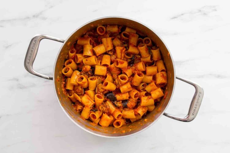 A stainless steel pot filled with rigatoni pasta mixed in a rich tomato pasta sauce, featuring small pieces of vegetables and tender eggplant, sits on a white marble surface.