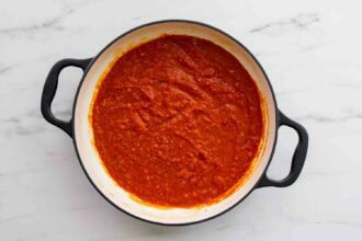 A round black-handled pot containing rich red tomato sauce for Pasta alla Norma sits on a white marble surface.