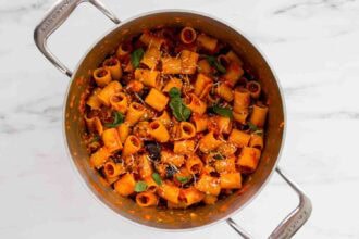 A pot of rigatoni tomato pasta mixed with sauce, garnished with fresh basil, grated cheese, and tender eggplant on a marble surface.