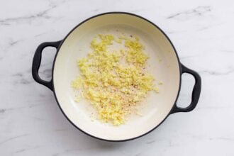 Chopped onions and seasonings are sautéing in a white pot with black handles on a marble countertop, forming the flavorful base for Pasta alla Norma or another delicious eggplant pasta dish.