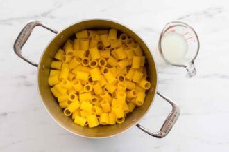A pot filled with uncooked pasta, perfect for making Pasta alla Norma, sits on a white surface next to a glass measuring cup containing a small amount of milk.