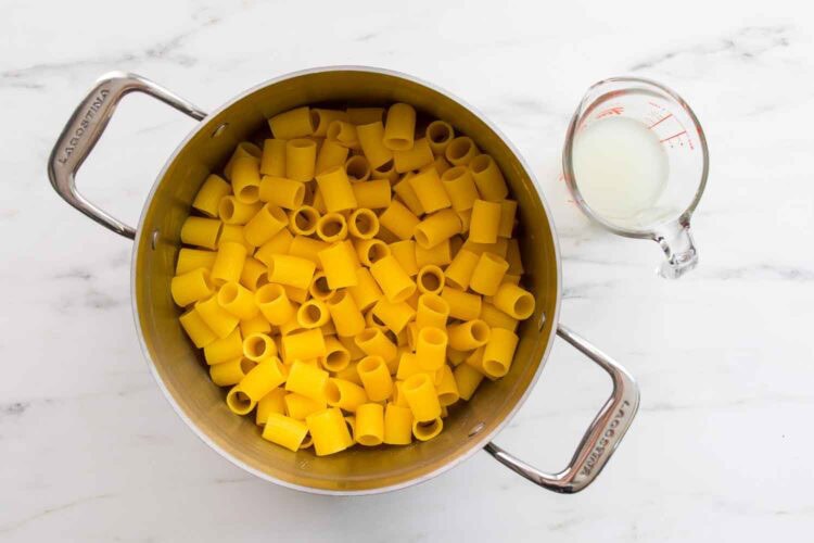 A pot filled with uncooked pasta, perfect for making Pasta alla Norma, sits on a white surface next to a glass measuring cup containing a small amount of milk.