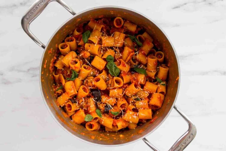 A pot of rigatoni tomato pasta mixed with sauce, garnished with fresh basil, grated cheese, and tender eggplant on a marble surface.