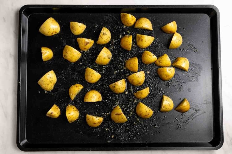 Quartered yellow potatoes coated in herbs and oil are evenly spread on a black sheet pan, ready to be roasted.