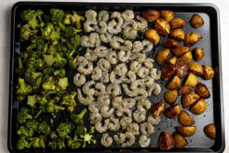 A sheet pan shrimp dinner featuring roasted broccoli florets on the left, seasoned shrimp in the center, and crispy roasted potato halves on the right.