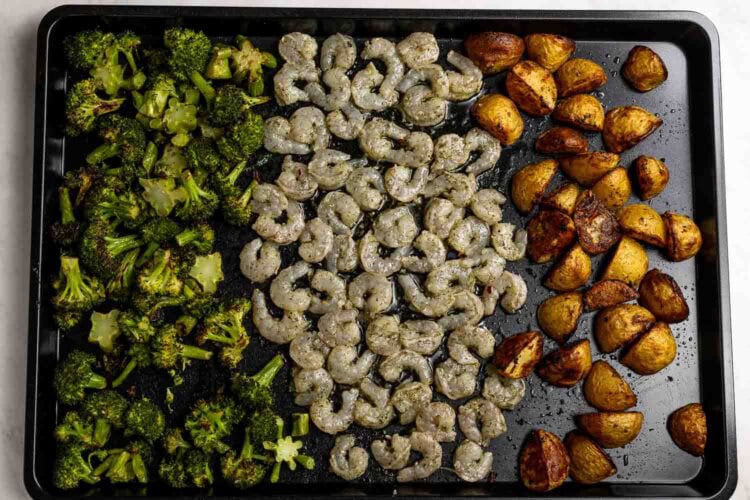 A sheet pan shrimp dinner featuring roasted broccoli florets on the left, seasoned shrimp in the center, and crispy roasted potato halves on the right.