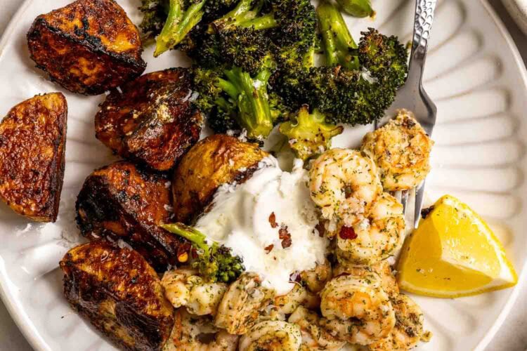 A plate with roasted potatoes, shrimp and broccoli in herbs, a dollop of sour cream, and a lemon wedge makes for a flavorful sheet pan dinner.