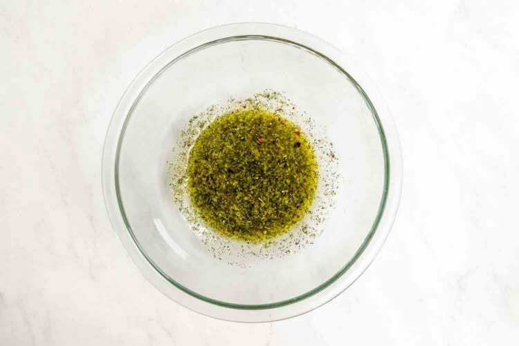 A clear glass bowl containing a mixture of green herbs and spices sits on a white surface, ready to add vibrant flavor to a delicious sheet pan shrimp and broccoli dinner.