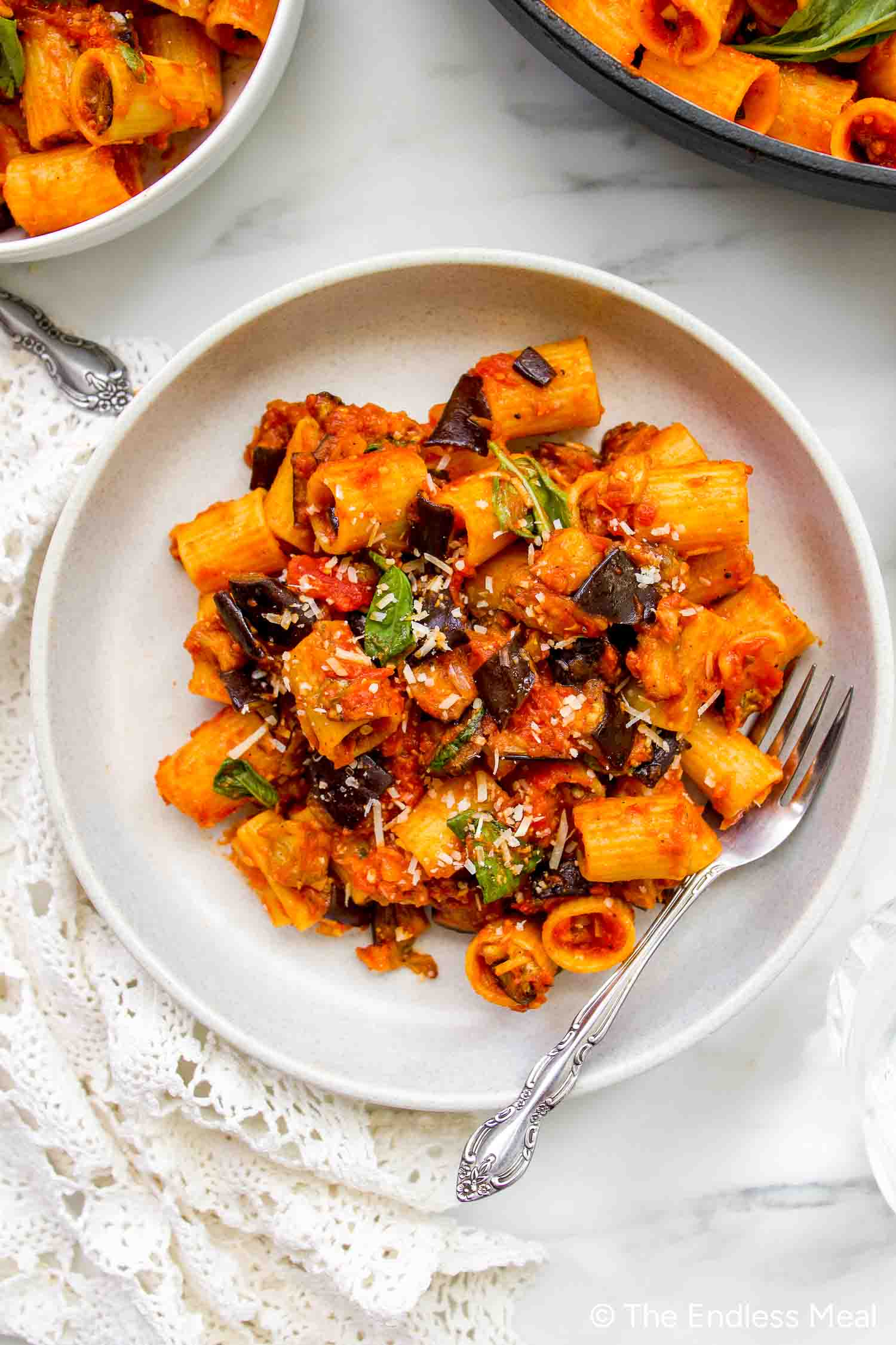 A plate of Pasta alla Norma features rigatoni tossed in tomato sauce with eggplant, basil, and grated cheese, served with a fork on a white dish.