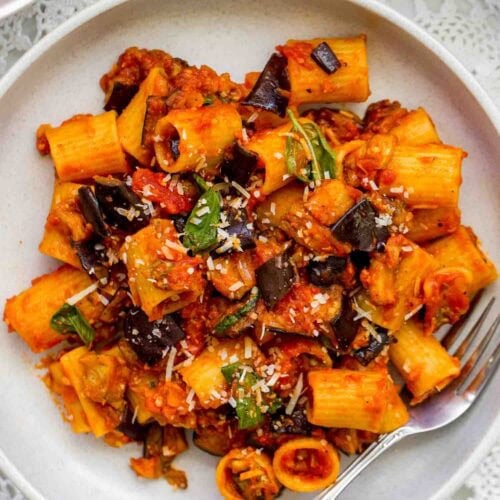 A plate of Pasta alla Norma with rigatoni, rich tomato sauce, eggplant, basil, and grated cheese is served with a fork on a white lace tablecloth.