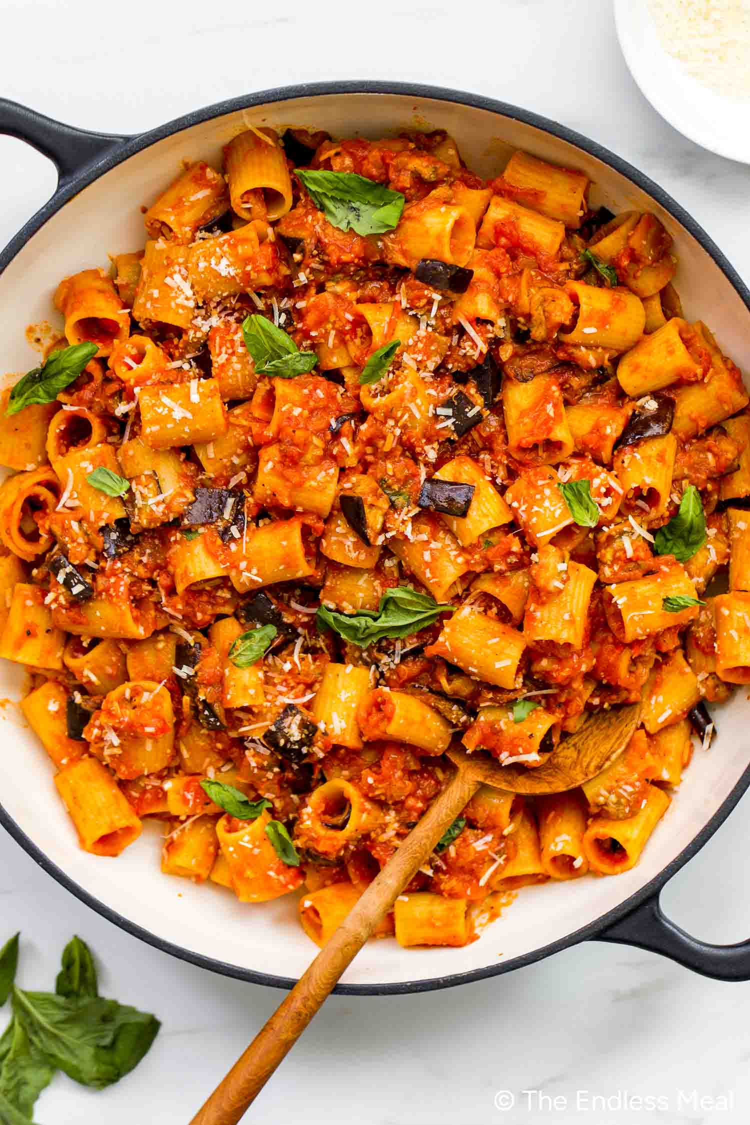 A pan filled with rigatoni in rich tomato pasta sauce, tender eggplant, fresh basil, and grated cheese—classic Pasta alla Norma—with a wooden spoon resting inside.