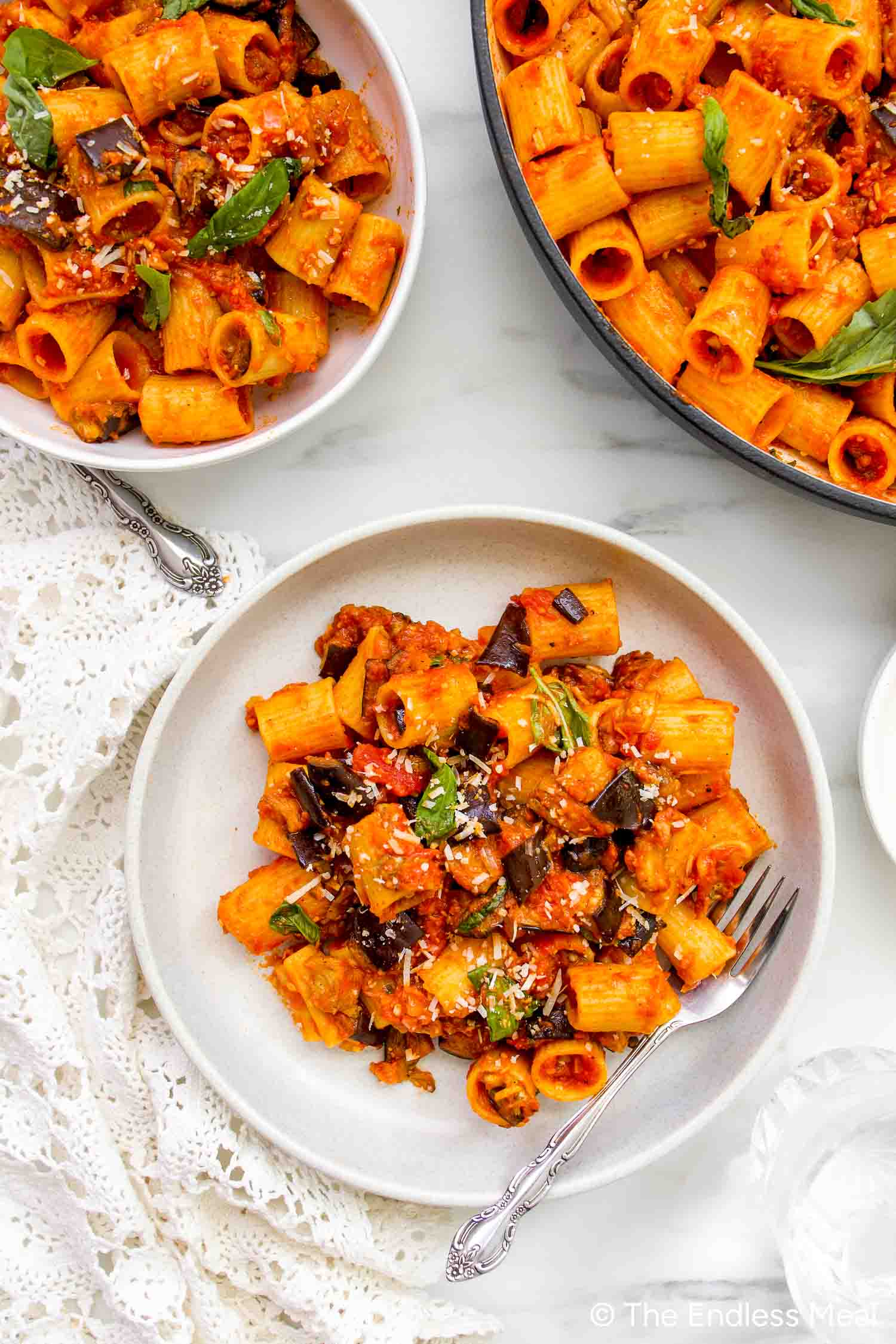 Plates of Pasta alla Norma, featuring rigatoni with tomato sauce, eggplant, and basil, topped with grated cheese, are set on a white surface near a black pan and a lace cloth.
