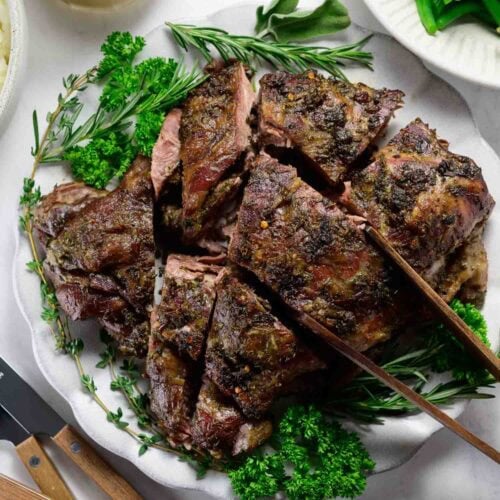 Sliced oven braised leg of lamb garnished with fresh herbs on a white plate, accompanied by green beans and a jug of gravy.