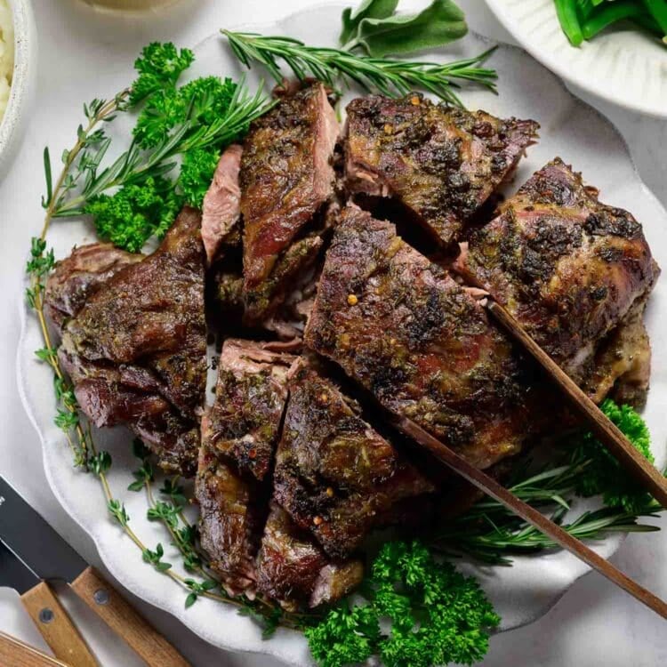 Sliced oven braised leg of lamb garnished with fresh herbs on a white plate, accompanied by green beans and a jug of gravy.