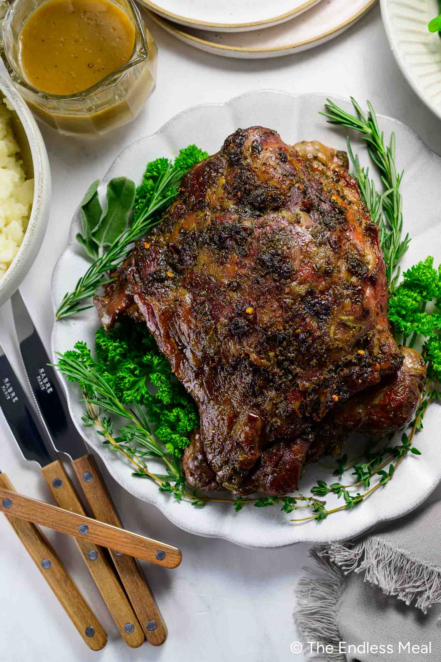 An Oven Braised Leg of Lamb, herb-crusted and cooked to perfection, sits surrounded by fresh green herbs on a white platter, with a bowl of gravy, mashed potatoes, and utensils nearby.
