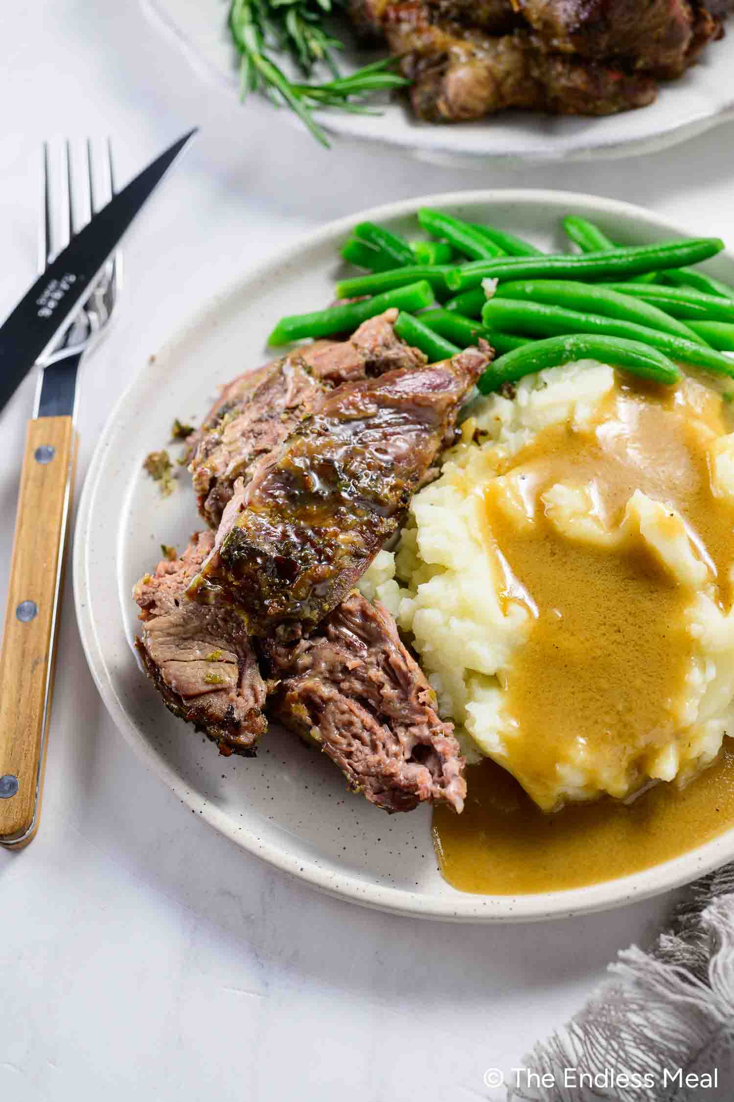 A plate with slices of roast beef, mashed potatoes topped with gravy, and green beans sits beside a fork and knife. Swap in boneless leg of lamb for a tender twist on this classic meal.