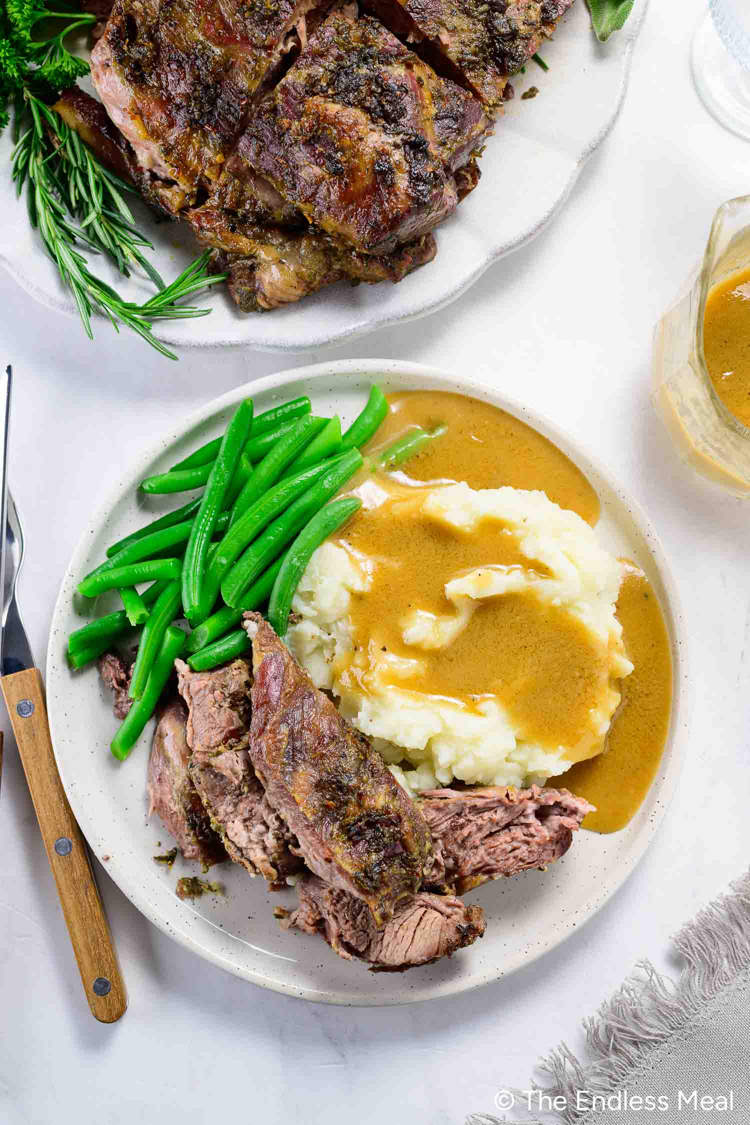 A plate with sliced boneless leg of lamb, mashed potatoes topped with gravy, and green beans. A fork and knife are beside the plate; extra meat and gravy are in the background.