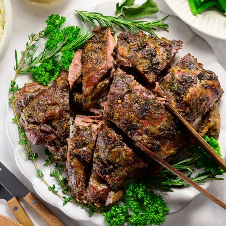 Sliced roasted lamb garnished with fresh herbs on a white plate, served with green beans and a jug of gravy nearby.