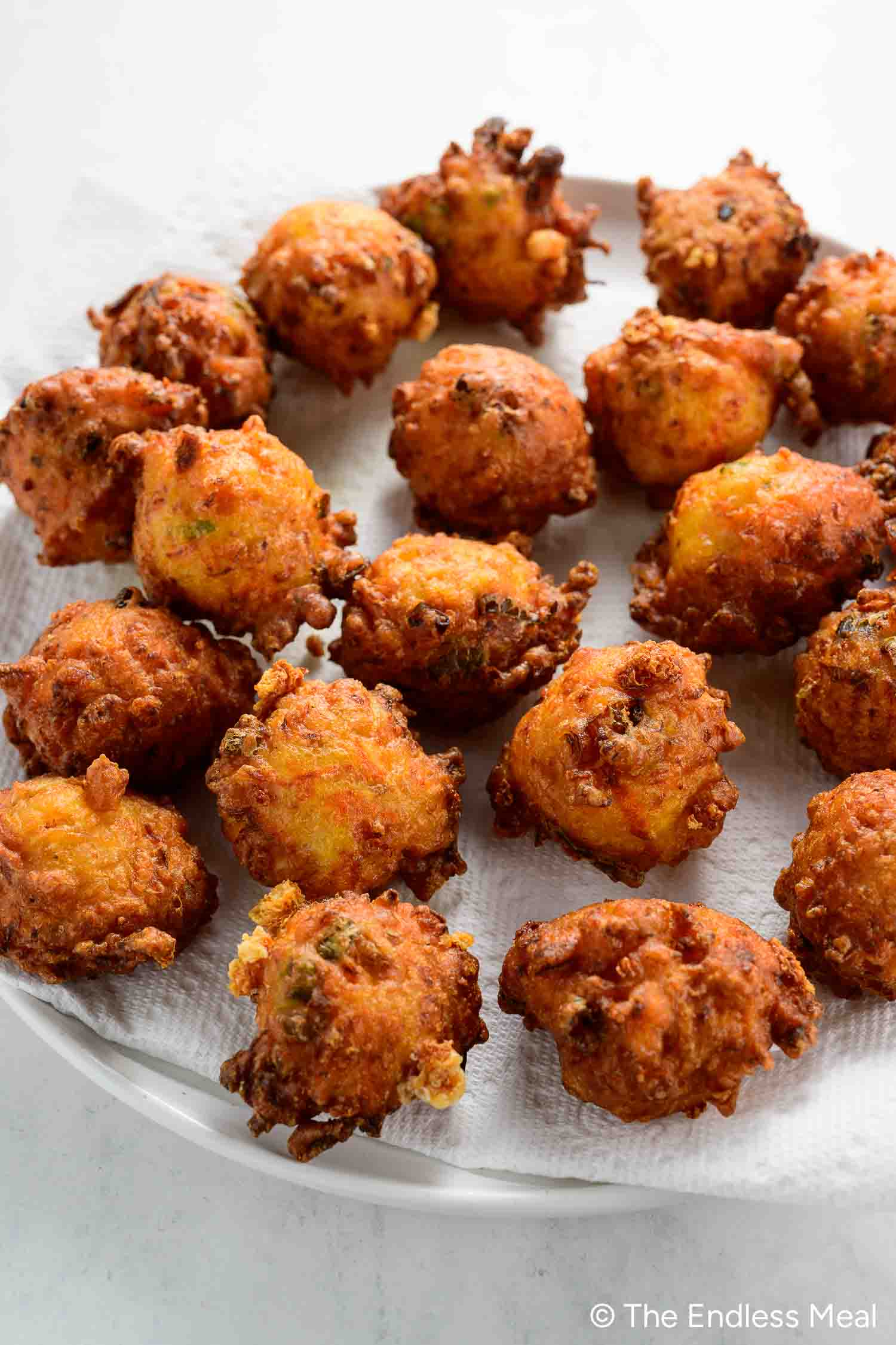 A plate of golden-brown vegetable fritters, reminiscent of classic hush puppies, sits on a paper towel arranged in a single layer.