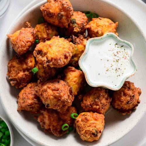 A bowl of golden-brown carrot fritters is served with a small dish of creamy dipping sauce and garnished with sliced green onions—a delicious twist on classic vegetable fritters.
