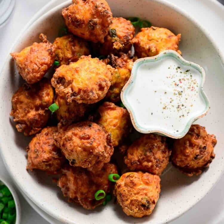 A bowl of golden-brown carrot fritters is served with a small dish of creamy dipping sauce and garnished with sliced green onions—a delicious twist on classic vegetable fritters.