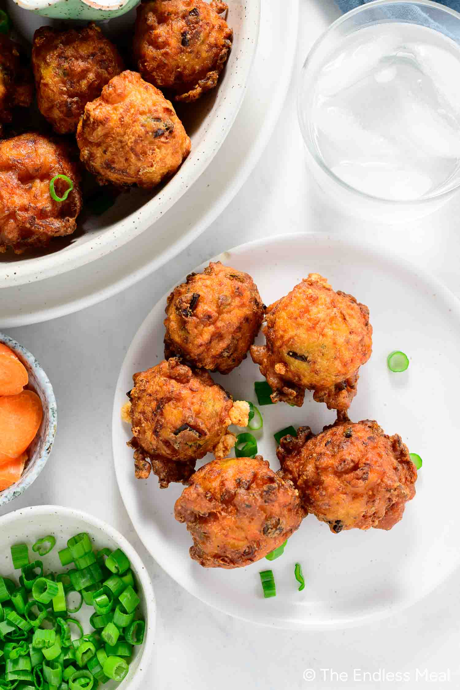 A plate with five golden-brown vegetable fritters, garnished with chopped green onions, next to bowls of more fritters, green onions, carrot slices, and a glass of water—a delicious choice for fans of Carrot Recipes.