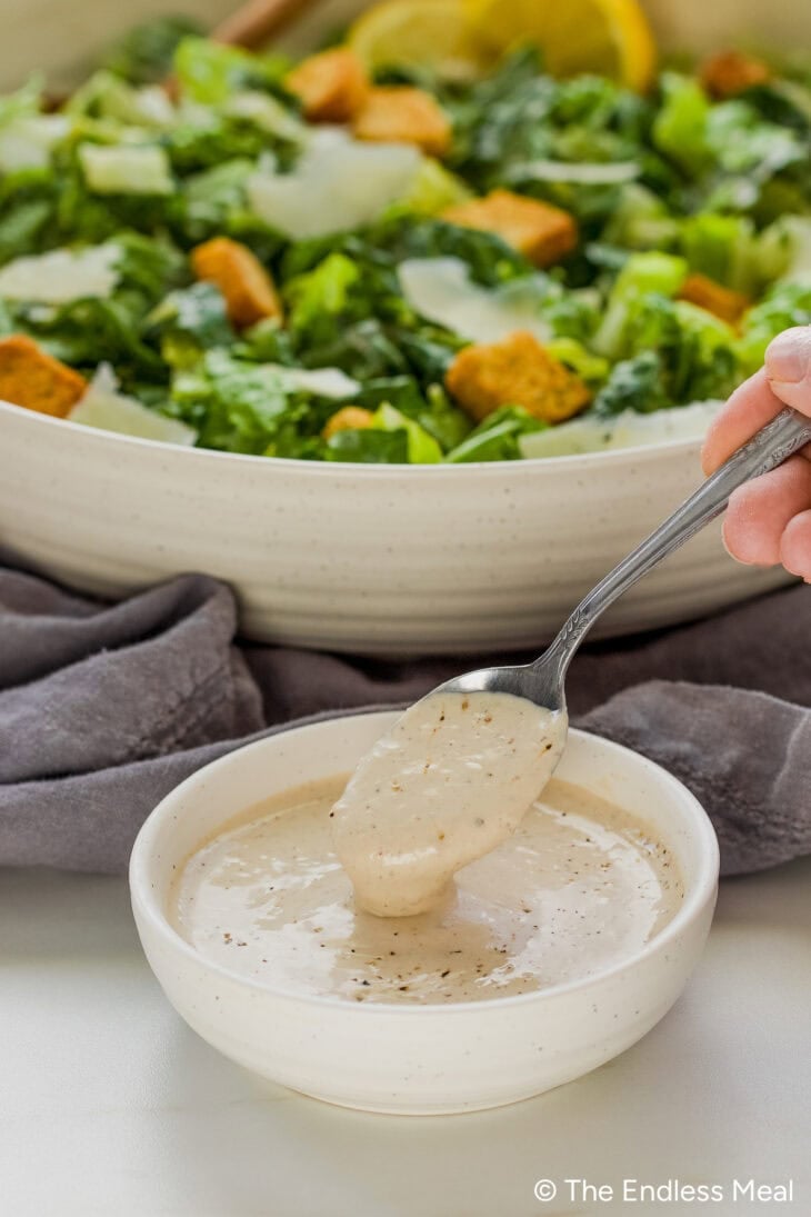 A spoon scoops creamy Greek Yogurt Caesar Salad Dressing from a small bowl, with a Caesar salad topped with croutons and Parmesan in the background.
