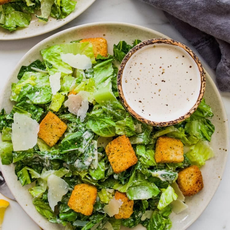A plate of Caesar salad with romaine lettuce, croutons, shaved parmesan, and a bowl of creamy Caesar Salad Dressing made with Greek Yogurt on the side for extra flavor.
