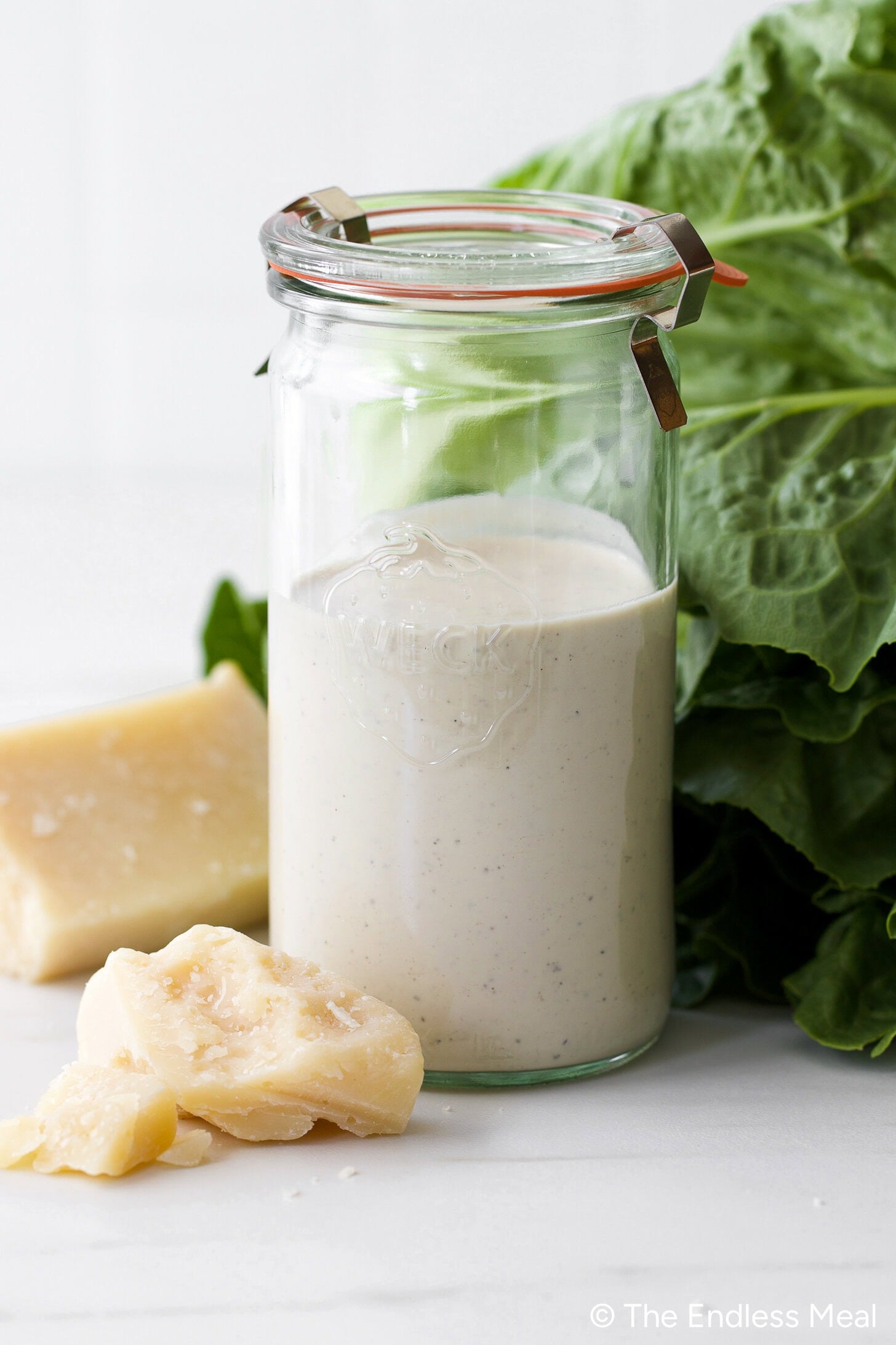 A glass jar filled with creamy Caesar Salad Dressing made with Greek Yogurt sits on a white surface, surrounded by leafy greens and chunks of Parmesan cheese, promising fresh flavor in every bite.