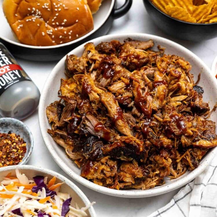 A plate of Hot Honey Pulled Pork with barbecue sauce, surrounded by sandwich buns, wavy potato chips, coleslaw, red pepper flakes, and a can of hard honey.