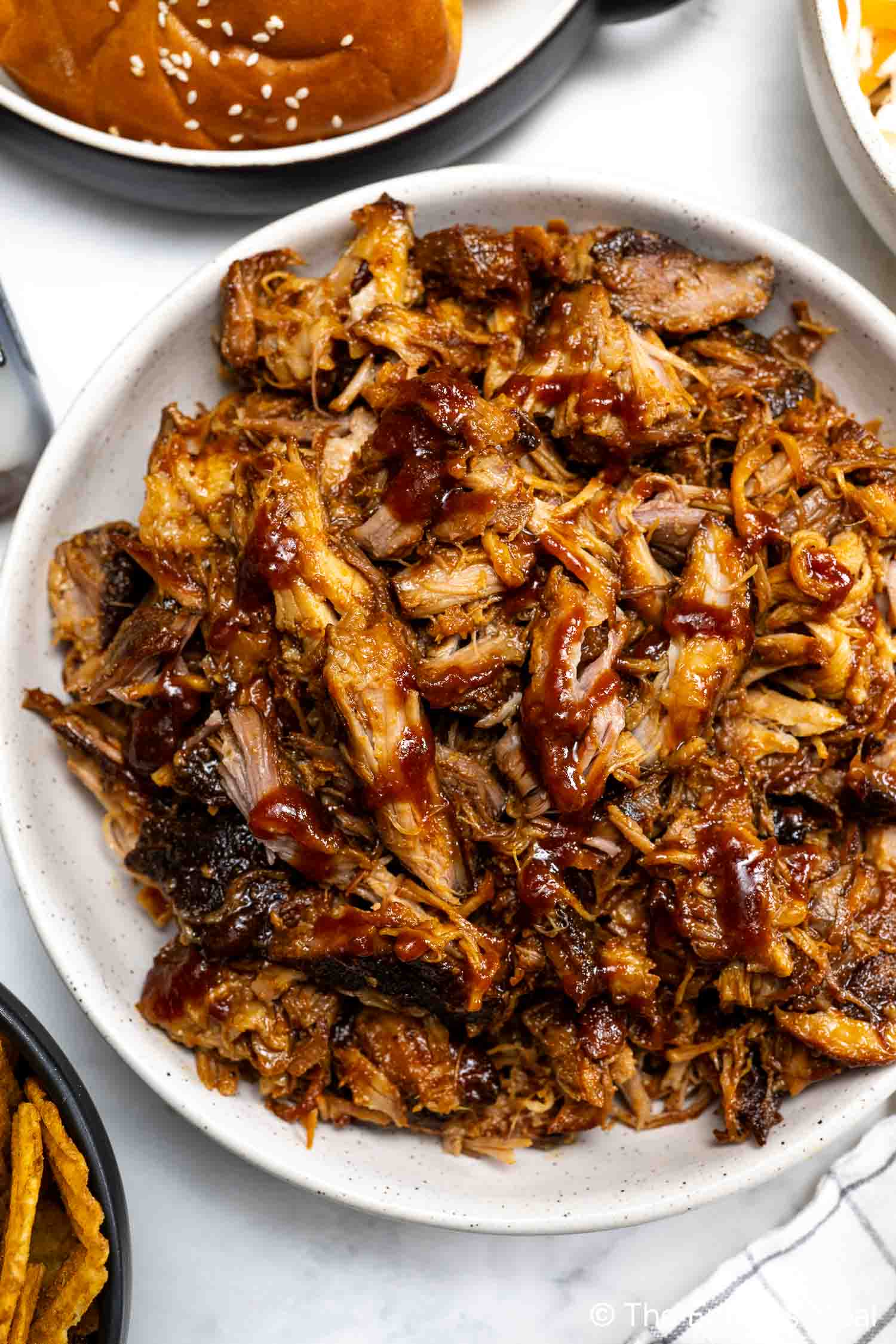 A white bowl filled with shredded Hot Honey Pulled Pork drizzled with barbecue sauce, with a sandwich bun partially visible in the background.